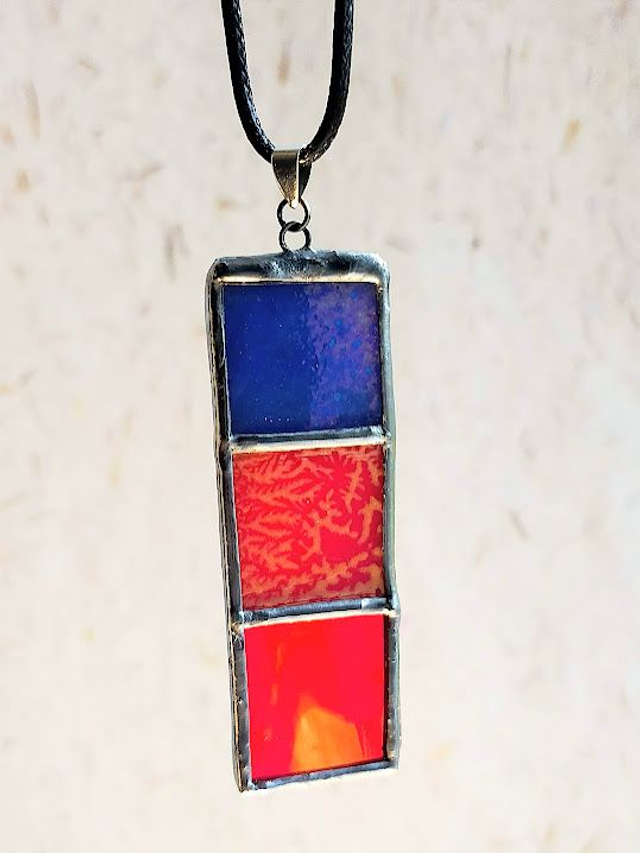 Pendentif en pâte de verre bleu, rouge et verre rouge- orangé, sertis en Tiffany et chaîne en coton ciré noir. 