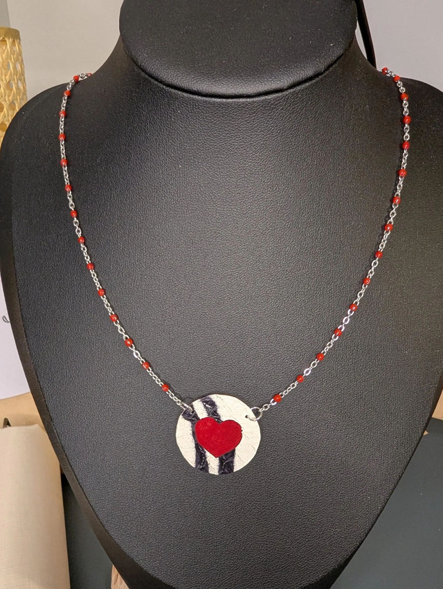 Collier mi long avec une chaine de couleur argent et rouge, avec un pendentif en cuir et en tissus enduit en forme de rond et de cœur. 