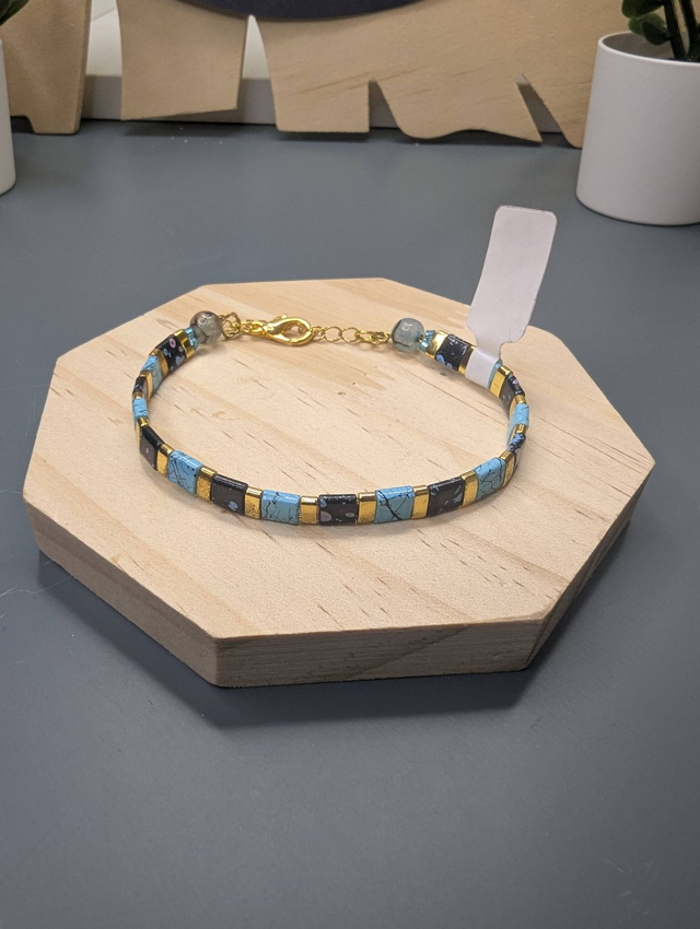 Bracelet avec des perles plate en verre noires et bleus et des perles en métal doré. Le fermoir et la chaine fermoir sont de couleur or.